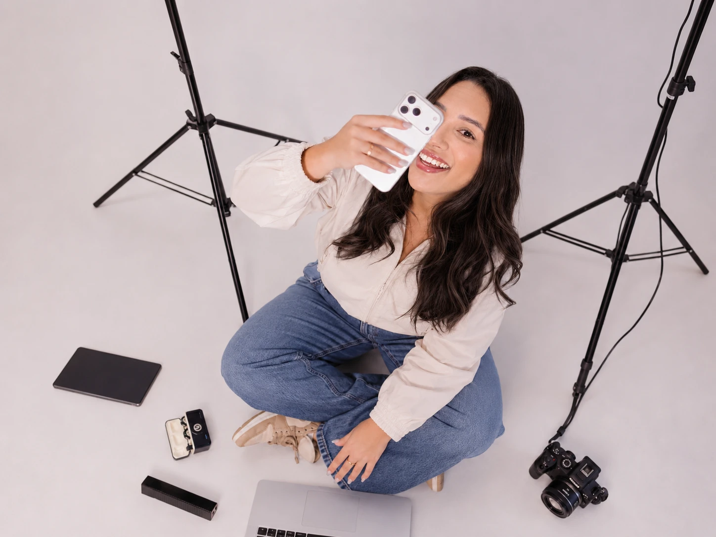 Mari Bonetto sentada no chão do estúdio entre tripés de luz, segurando o celular perto do rosto e sorrindo, com câmera, notebook e microfone ao redor