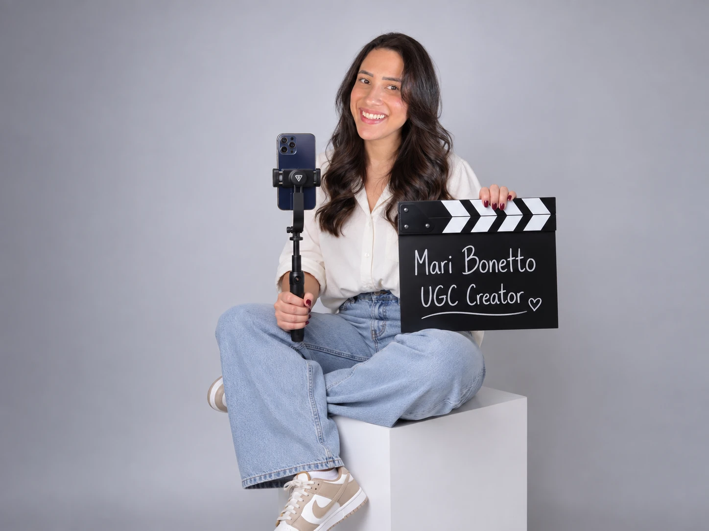 Mari Bonetto sentada em um cubo branco, sorrindo, segurando um celular no estabilizador e uma claquete escrita Mari Bonetto UGC Creator